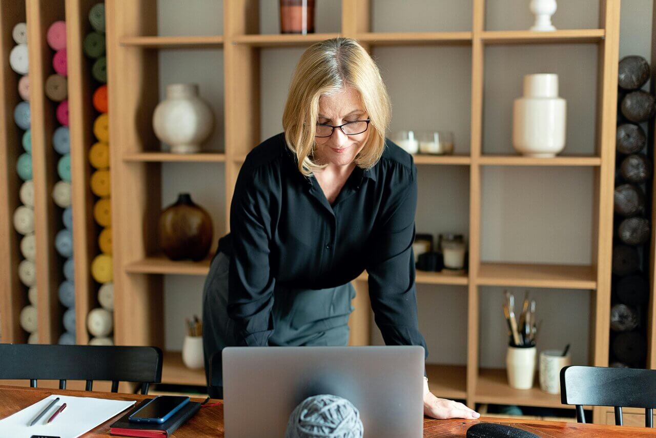 Older blonde woman standing over desk looking at laptop 